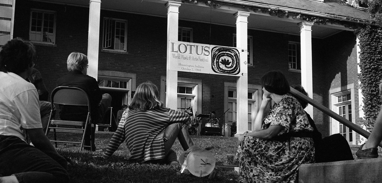 Image of a group of people sitting on lawn of Museum's administrative annex listening to a band performing under a "Lotus Festival" banner.