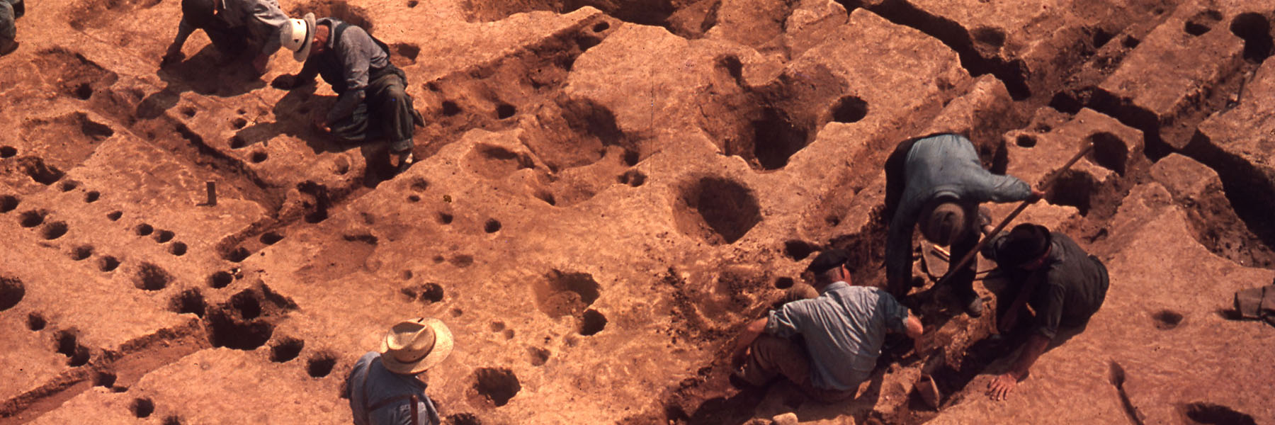 Six men looking at multiple circular holes in the ground at an excavation site.